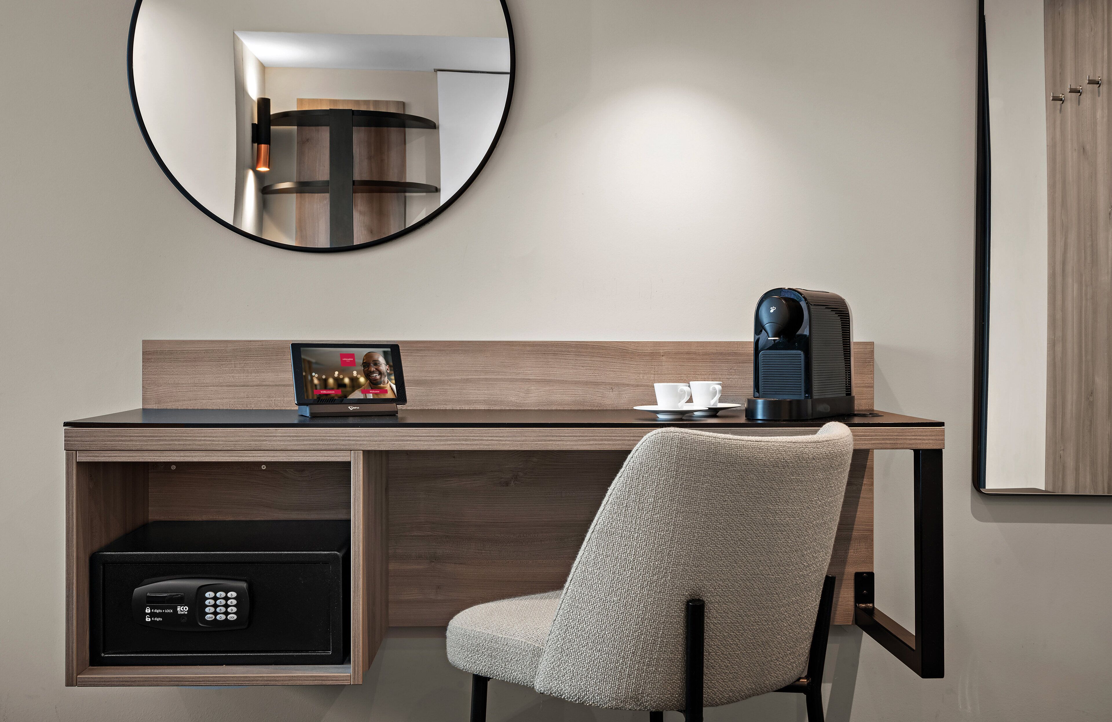 A modern hotel room with a wooden desk, a chair, a coffee maker, a framed picture, and a safe.