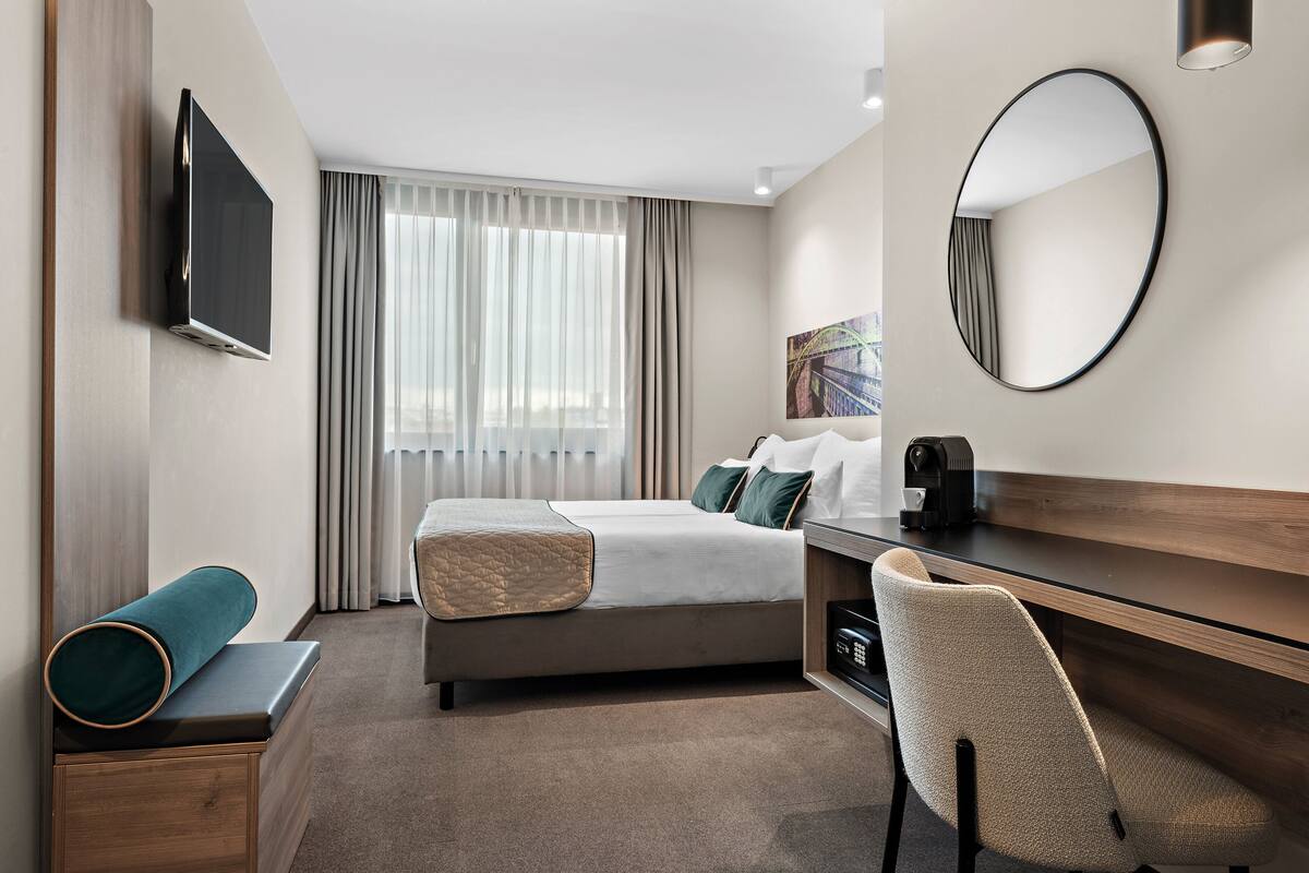 A modern hotel room with a bed, a desk, a chair, a TV, a mirror, and a window with curtains.