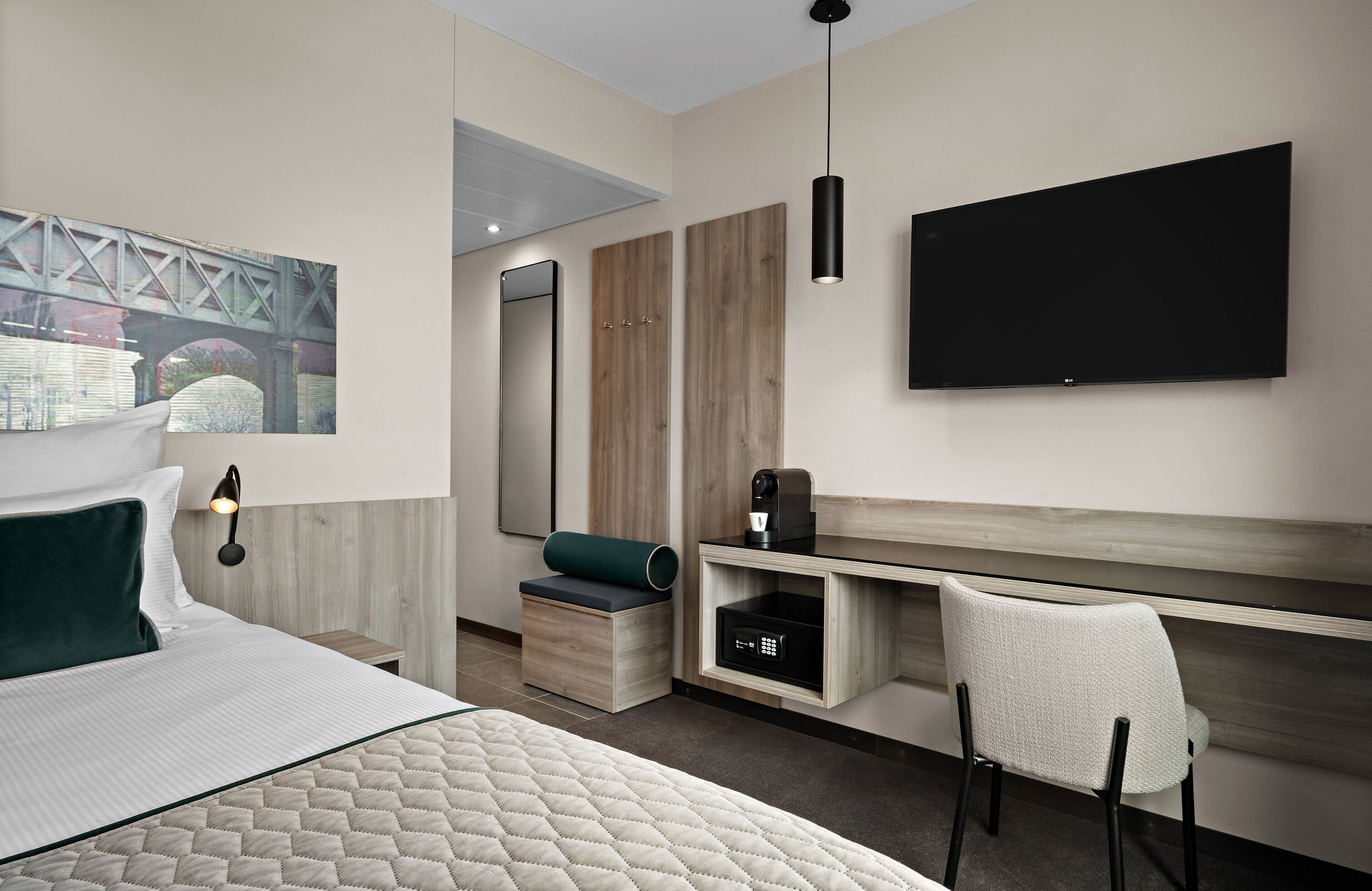 A modern hotel room with a bed, a desk, a chair, a flat-screen TV, a coffee machine, and a painting on the wall.