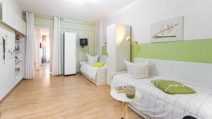 Una habitación moderna y bien iluminada con piso de madera, una cama con almohada verde, un sofá blanco con almohada verde, una mesita con un libro y una lámpara, y un cuadro en la pared.