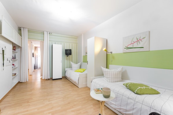 Une pièce moderne et bien éclairée, avec un plancher en bois, un lit avec un oreiller vert, un canapé blanc avec un oreiller vert, une petite table avec un livre et une lampe, et un tableau accroché au mur.