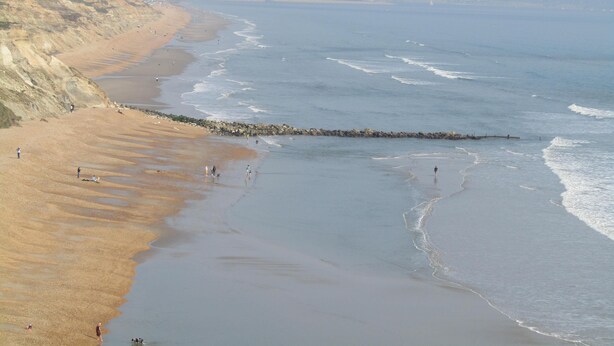 Uma praia costeira com um molhe rochoso, areal arenoso e mar calmo.