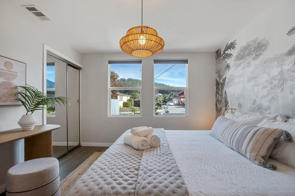 Un dormitorio con una cama grande, una lámpara colgante, vistas a través de la ventana y una planta sobre una mesita de noche.