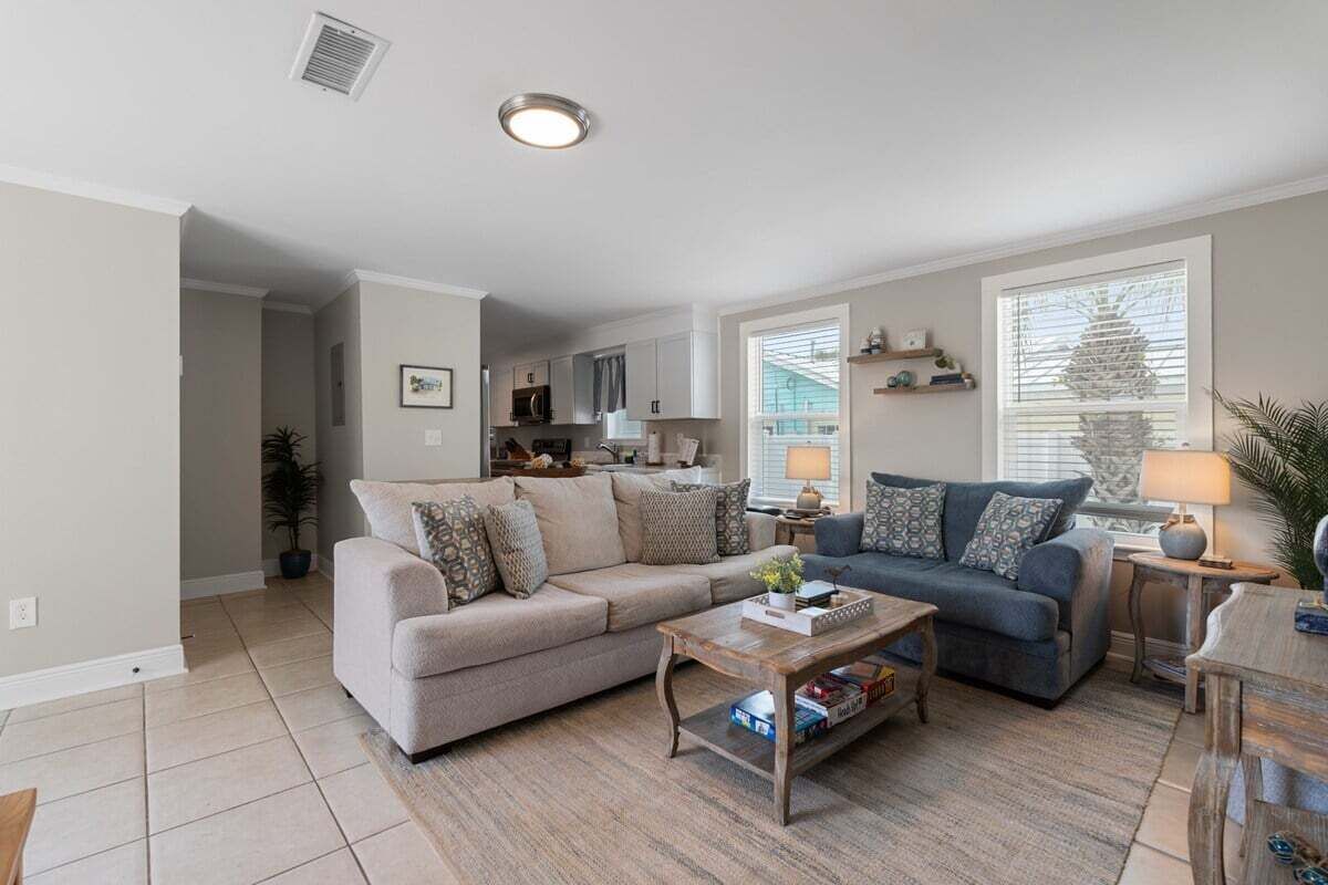A living room with a beige sofa, a blue sofa, and a wooden coffee table. There is a kitchen in the background with white cabinets and a window with blinds.