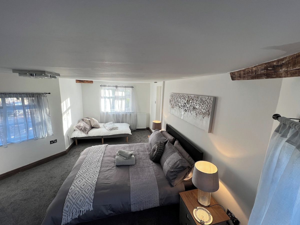 A bedroom with a large bed, a smaller bed, a bedside table with a lamp, and a window with curtains.
