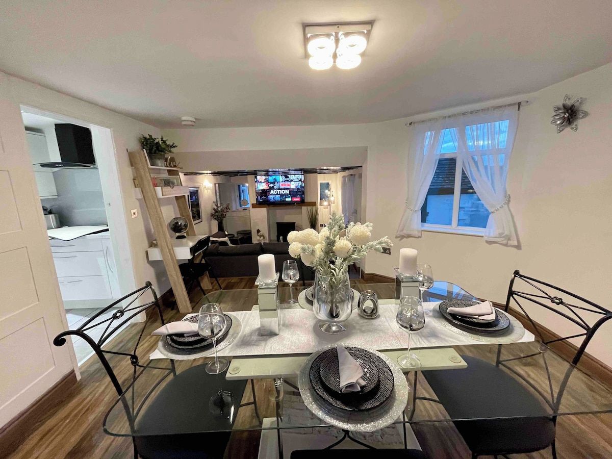 A dining area with a table set for four, a television in the background, and a window with curtains.