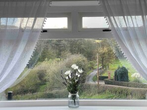 A window with sheer curtains, a vase with flowers, and a view of a landscaped garden.