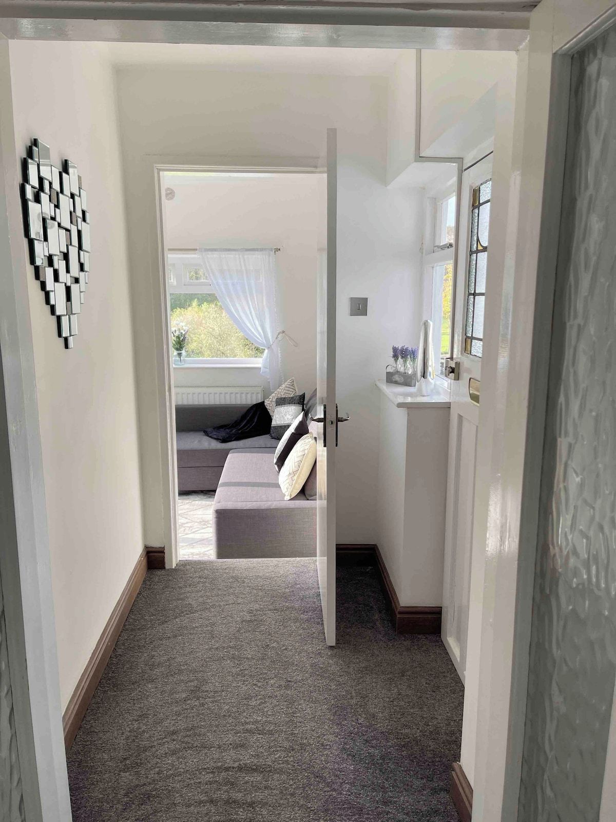 A hallway leading to a bedroom with a bed, a window with curtains, and a wall-mounted mirror.