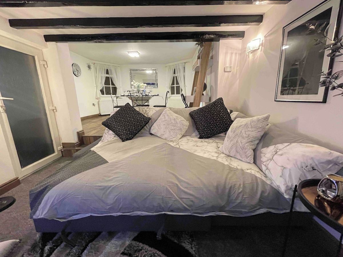 A bedroom with a large bed, a bedside table, a mirror, and a view into the dining area.