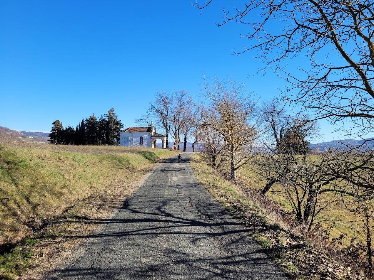 A rural road leading to a building with a red roof and a bell tower, surrounded by trees and open fields.