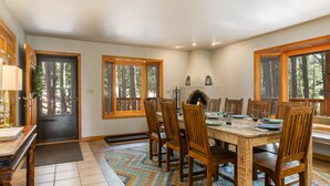 A dining area with a wooden table, chairs, a fireplace, and large windows with a forest view. - Cabin Walkable to Lake with Hot Tub, Deck & Views, Pool Table, Near Golf & Bike (Angel Fire)