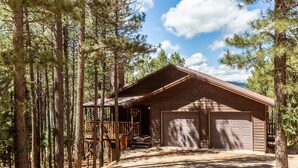 A cabin with a wooden deck, a garage, and surrounded by tall pine trees. - Cabin Walkable to Lake with Hot Tub, Deck & Views, Pool Table, Near Golf & Bike (Angel Fire)