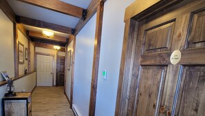 Un pasillo con piso de madera, una alfombra negra en la entrada, una puerta de madera con una placa identificativa y una lámpara en el techo.