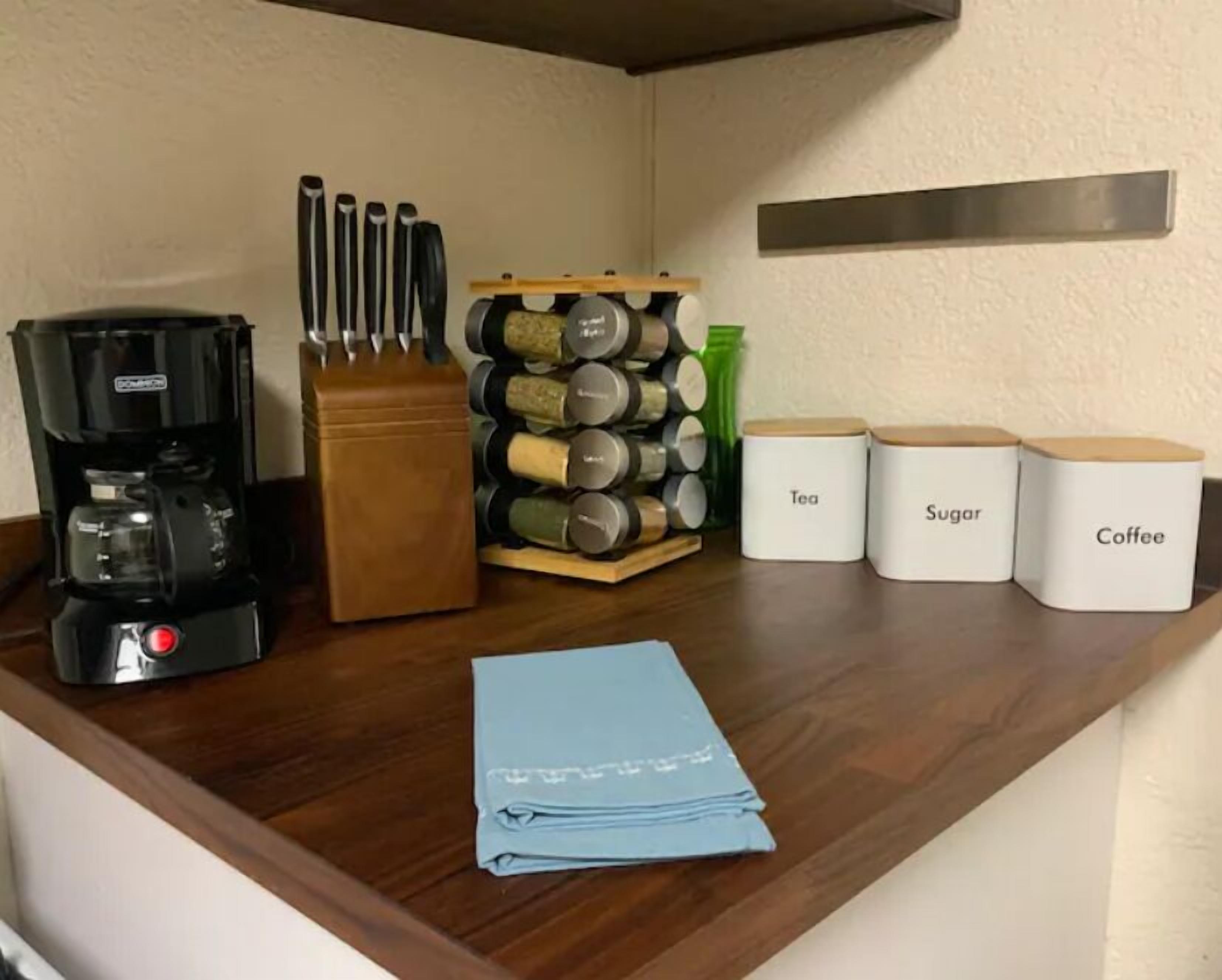 Un plan de travail de cuisine avec une machine à café, un bloc à couteaux, des pots à épices et des récipients étiquetés pour le thé, le sucre et le café.