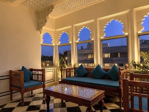 A room with a checkered floor, a wooden sofa with teal cushions, a wooden coffee table, and a window with decorative cutouts. - The Amar Mahal (Udaipur)