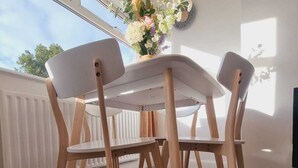Un comedor con una mesa y sillas blancas, un jarrón de flores y vistas al exterior a través de amplias ventanas.