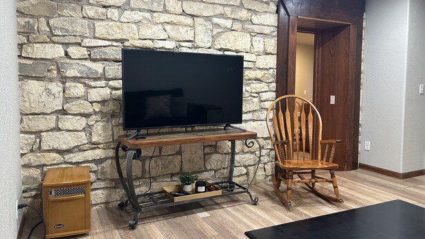 A living room with a stone wall, a flat-screen TV on a wooden stand, a rocking chair, a small heater, and a wooden coffee table. - Unique Space in Historic Building (Osborne)