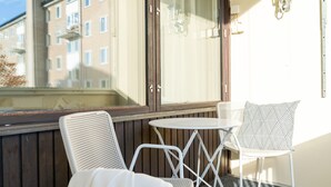 En balkong med två vita metallstolar och ett litet runt bord, träpanel och utsikt över en byggnad med många fönster.