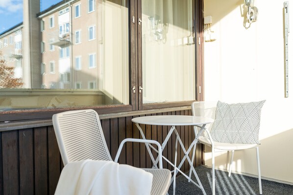 A balcony with two white metal chairs and a small round table, wooden paneling, and a view of a building with multiple windows.