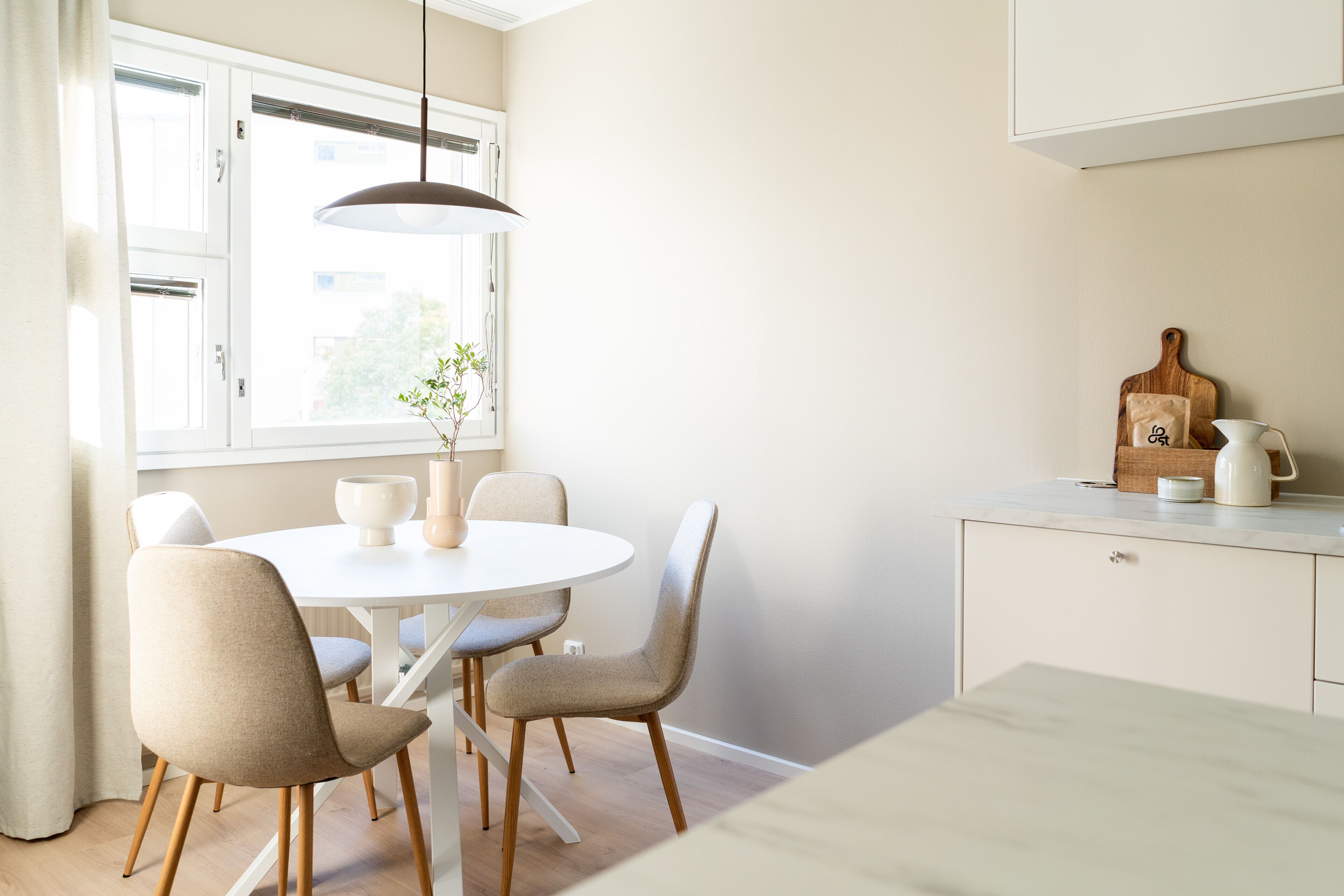 A dining area with a round table, four chairs, a hanging light fixture, a window with curtains, and a kitchen counter with a cutting board and a kettle.