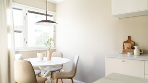 En matplats med ett runt bord, fyra stolar, en hängande lampa, ett fönster med gardiner och en köksbänk med en skärbräda och en vattenkokare.