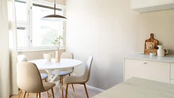 A dining area with a round table, four chairs, a hanging light fixture, a window with curtains, and a kitchen counter with a cutting board and a kettle.