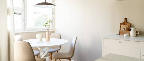 A dining area with a round table, four chairs, a hanging light fixture, a window with curtains, and a kitchen counter with a cutting board and a kettle.