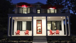 Una casa de dos pisos con un porche cubierto, una puerta roja y dos sillas rojas.