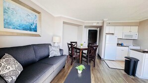 Uma sala de estar moderna com um sofá cinzento, uma mesa de centro e um espaço de refeições com cadeiras de madeira. Uma cozinha com armários e eletrodomésticos brancos é visível ao fundo.