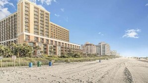 Um hotel à beira-mar com vários andares, uma praia de areia em primeiro plano e outros edifícios ao fundo.