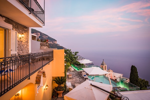 A coastal hotel with a balcony overlooking the sea, outdoor seating, and a view of a church.