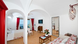 Un salon spacieux avec un canapé, une petite table et un fauteuil. Il y a un balcon avec des rideaux rouges et une vue sur l’extérieur.