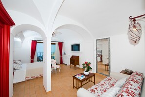 A spacious living room with a sofa, a small table, and a chair. There is a balcony with red curtains and a view of the outside.