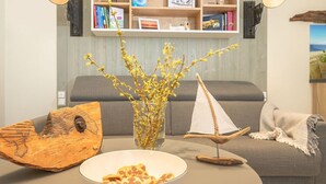 Ein Wohnzimmer mit grauem Sofa, einer Glaskaraffe mit gelben Blumen, einem hölzernen Schiffsmodell und einem Teller mit Essen.