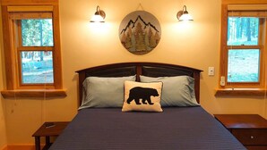 A bedroom with a bed, two bedside lamps, a wall-mounted clock, and windows with a view of trees.