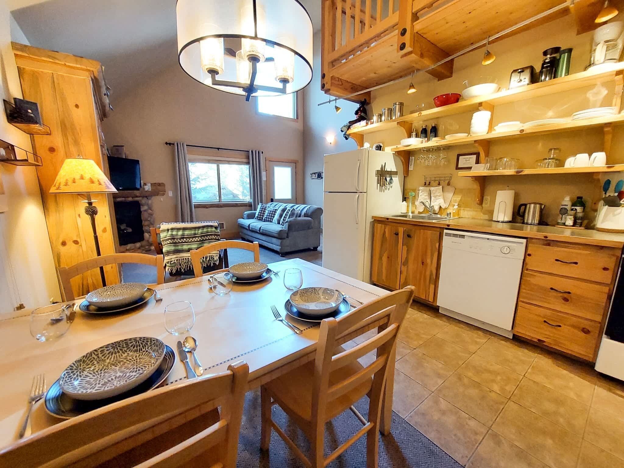 Una cocina acogedora con armarios de madera, una mesa de comedor preparada para cuatro personas y una sala de estar con un sofá y un televisor.