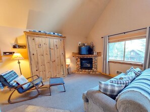 A cozy living room with a stone fireplace, wooden cabinets, a television, and a large window. - Alpine View Cabin - duplex unit B w/ queen bedroom (Winthrop)