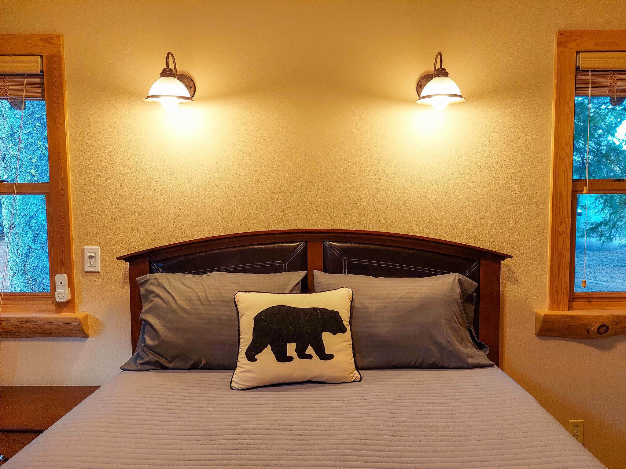 Una cama con cabecera, dos lámparas de pared, una ventana con vista a árboles y un enchufe.