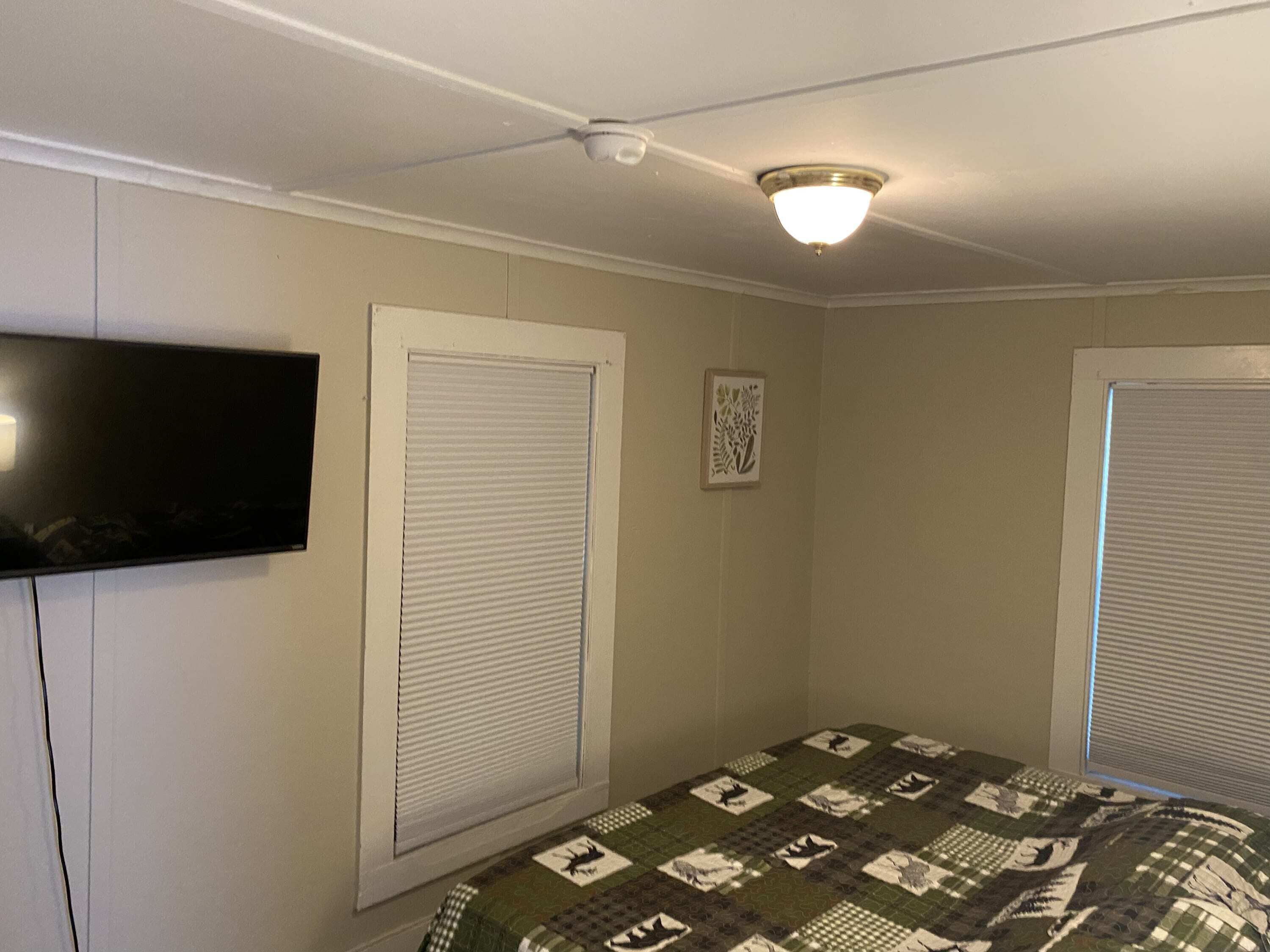Une chambre avec deux fenêtres, un ventilateur de plafond, un téléviseur à écran plat fixé au mur et une courtepointe sur le lit.