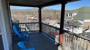 Un porche cubierto con sillas Adirondack azules y una pequeña mesa, con vistas a una zona residencial con casas y montañas al fondo.