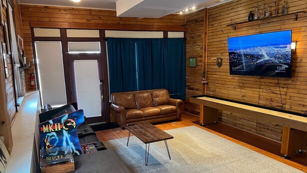 Una sala de estar acogedora con un sofá de cuero marrón, una mesa de centro de madera y una mesa de billar. Un televisor montado en la pared muestra un paisaje urbano.