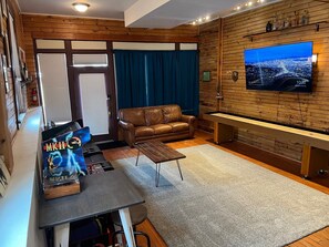 A cozy living room with a brown leather sofa, a wooden coffee table, and a pool table. A television mounted on the wall displays a cityscape.
