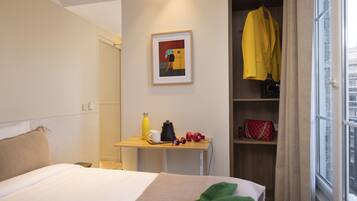 Ein Hotelzimmer mit einem Bett, einem Schreibtisch mit Wasserkocher, einer roten Tasche und einer gelben Jacke, die an der Wand hängt.