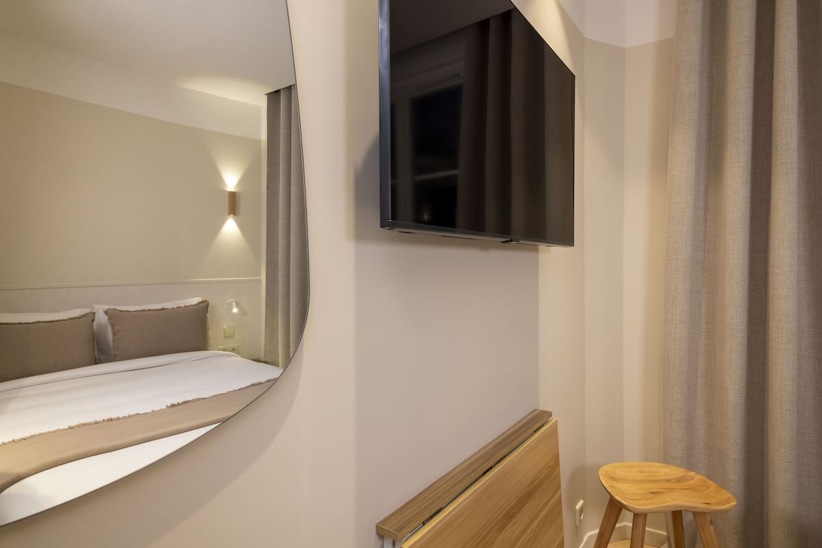 Ein Hotelzimmer mit einem Bett, einem an der Wand befestigten Fernseher, einem Nachttisch aus Holz und einem Fenster mit Vorhängen.