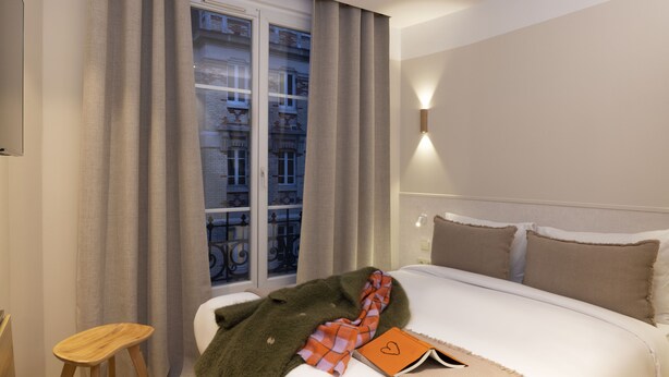A hotel room with a bed, a window with curtains, a bedside table, and a wall-mounted light. - Le Daum (Paris)