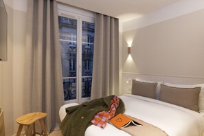 A hotel room with a bed, a window with curtains, a bedside table, and a wall-mounted light. - Le Daum (Paris)