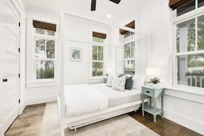 Quarto com cama branca, mesa de cabeceira e janela com vista para árvores.