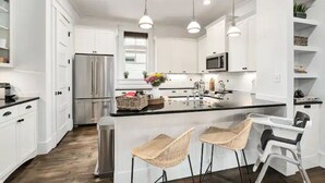 A modern kitchen with white cabinetry, stainless steel appliances, a central island with bar stools, pendant lighting, and a window providing natural light. - Kid friendly home with golf cart, sports gear for beach pool lake  (Inlet Beach)