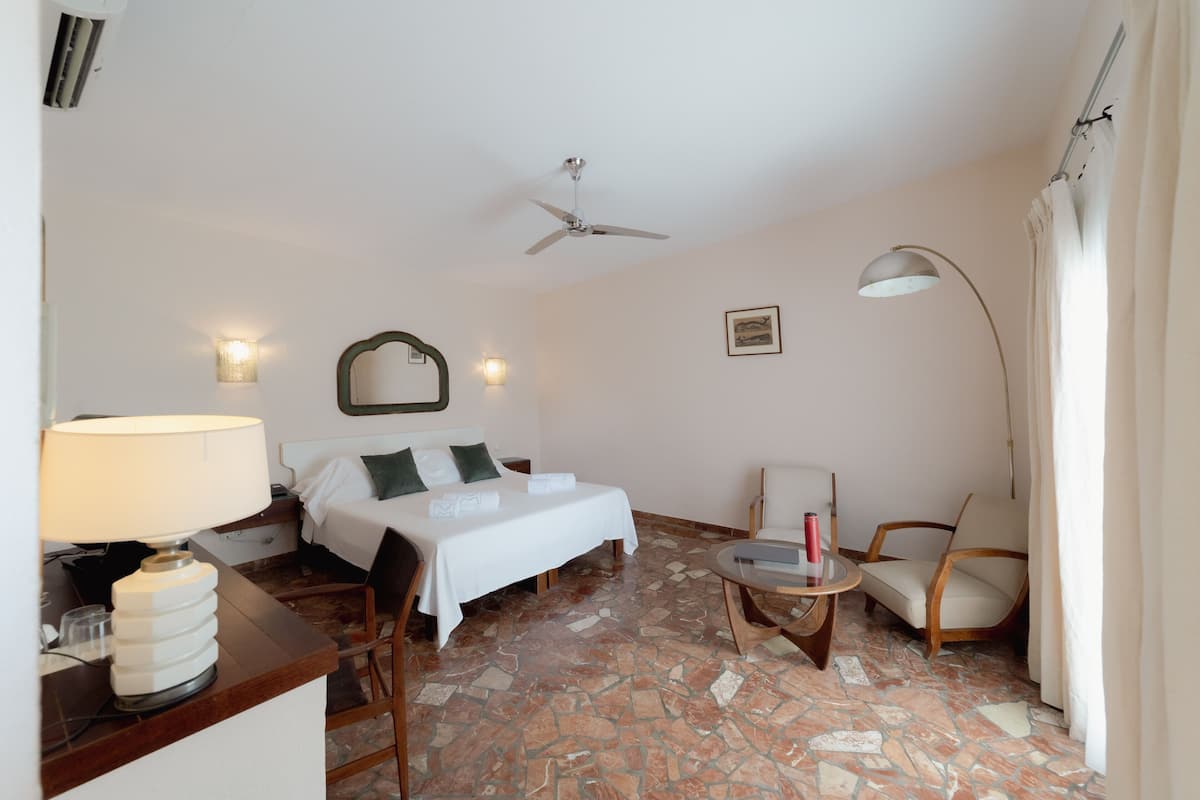 A hotel room with a bed, a desk with a lamp, a chair, a small table, a ceiling fan, and a floor lamp.