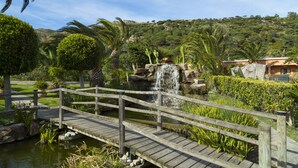 A wooden bridge over a pond with a waterfall, surrounded by lush greenery and a hillside in the background. - Hotel Punta Sur (Tarifa)